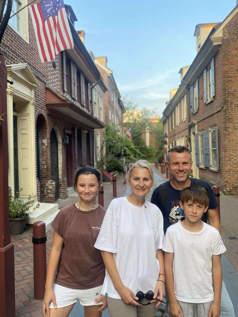 Une famille de quatre personnes pose dans une rue historique pavée de briques, bordée de vieux bâtiments en briques et de drapeaux américains, sous un ciel bleu et clair. Ils sont souriants.