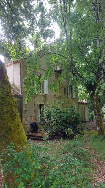 Ein charmantes altes Haus mit beigen Wänden und weißen Fensterläden liegt eingebettet zwischen üppigen, grünen Bäumen. Moos und Laub sorgen für eine ruhige, natürliche Atmosphäre.