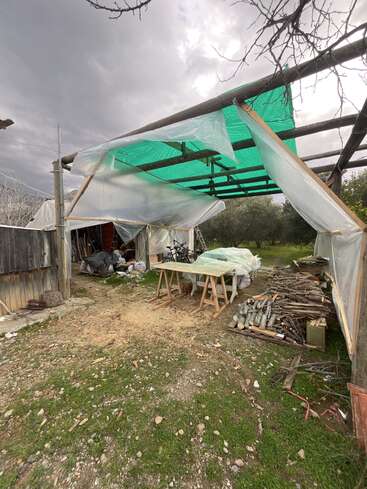 Un espace de travail rustique en plein air avec un toit de fortune recouvert de plastique, du bois de chauffage empilé, des tréteaux, des matériaux éparpillés, des outils et un ciel nuageux au-dessus, suggérant des projets récents ou en cours.