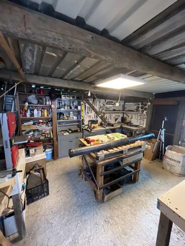 Un atelier bien organisé avec des étagères remplies d'outils, de matériel et de matériaux. Un grand établi trône au centre, sur lequel sont disposés divers outils et pièces.