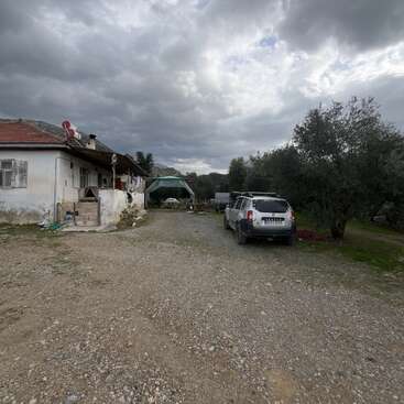 Une maison rustique au toit rouge se trouve dans une cour gravillonnée sous un ciel nuageux. On aperçoit un SUV blanc, des arbres et un espace extérieur ombragé.