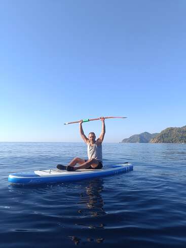 Ein Typ sitzt auf einem Paddleboard im ruhigen blauen Wasser und hält triumphierend ein Paddel über seinen Kopf, mit malerischen Bergen und klarem Himmel im Hintergrund.