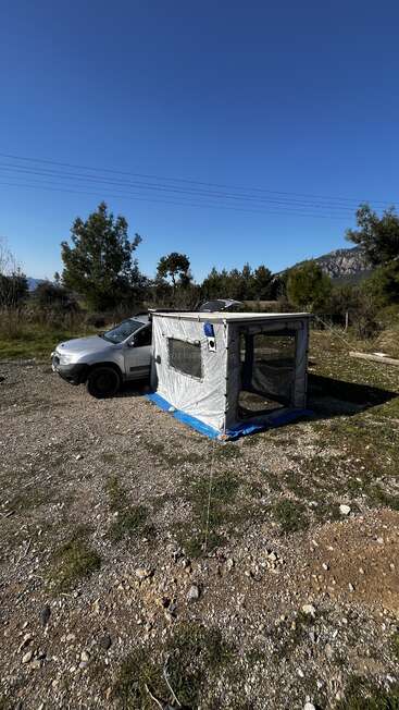 Ein silbernes Auto steht auf felsigem Boden und ist an einem kleinen grauen Zelt oder Vordach befestigt. Bäume, Berge und ein klarer blauer Himmel machen den Hintergrund komplett.