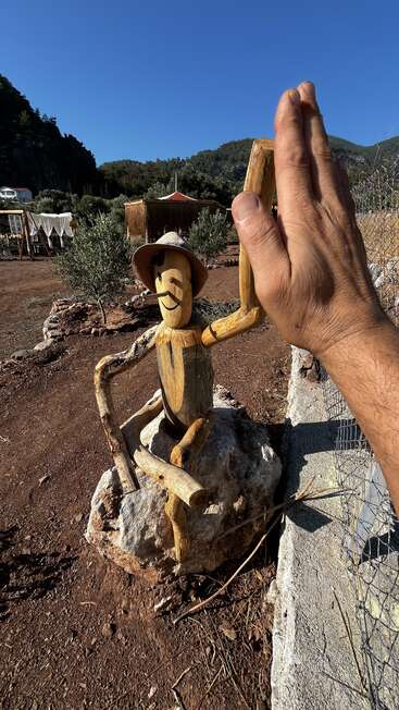 Eine Holzfigur mit Hut und lächelndem Gesicht sitzt auf einem Felsen. Jemand gibt ihr unter strahlend blauem Himmel ein High Five.