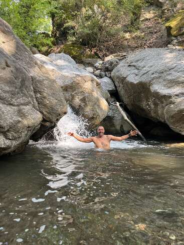 Ein lächelnder Mann badet fröhlich in einem klaren, felsigen Bach, die Arme ausgestreckt, umgeben von großen Felsbrocken und üppigem, grünem Wald im Sonnenlicht.