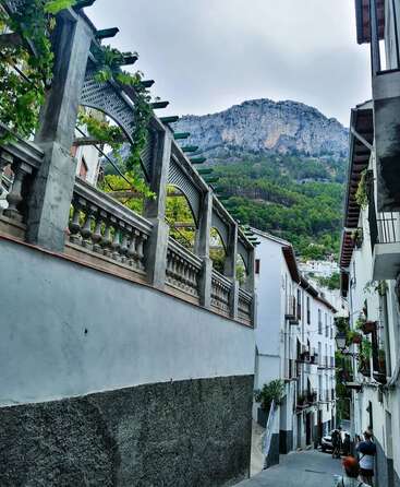 Eine schmale Straße schlängelt sich zwischen weiß getünchten Häusern, üppigem Grün entlang einer Terrasse, unter der Menschen spazieren gehen und einem dramatischen Berg, der sich majestätisch im Hintergrund erhebt.