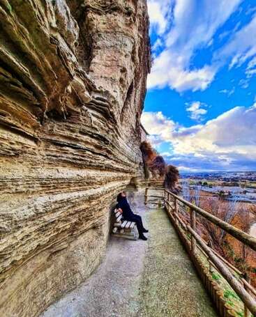 Eine Person entspannt sich auf einer in den Fels gehauenen Bank mit Blick auf ein malerisches Tal unter einem strahlend blauen Himmel mit dramatischen, verstreuten Wolken. Gelassene Atmosphäre.