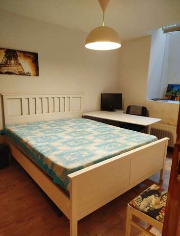 Ein gemütliches Schlafzimmer mit einem großen weißen Bett, einer blau gemusterten Decke, einem Schreibtisch mit Computer, einem Stuhl, Wandkunst und einer Hängelampe über dem Kopf sowie einem Holzfußboden.