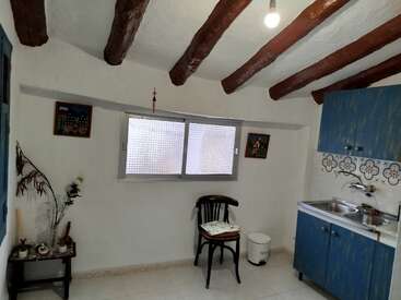 Essa cozinha aconchegante apresenta vigas de madeira expostas no teto, um pequeno armário azul, uma cadeira simples, obras de arte em paredes brancas e uma janela que fornece luz natural.