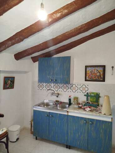 Esta é uma pequena cozinha rústica com vigas de madeira expostas no teto, armários azuis, um backsplash de azulejos, uma pia dupla, arte na parede e iluminação mínima.