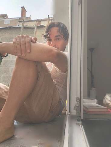 Um homem senta-se descalço em uma janela iluminada pelo sol, abraçando os joelhos. Ele olha para fora, relaxado. Você pode ver livros, uma lâmpada e um ambiente interno suave.