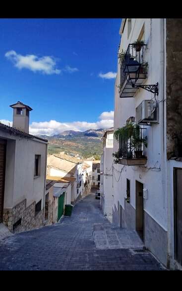 A narrow cobblestone street winds through a quaint village with white-washed buildings, balconies with plants, and mountain scenery beneath a bright blue sky. Peaceful atmosphere.