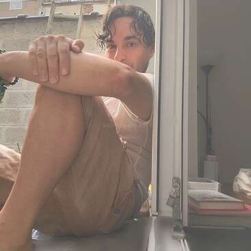 Ein Mann sitzt barfuß vor einem sonnenbeschienenen Fenster und stützt sich auf die Knie. Er blickt entspannt nach draußen. Bücher, eine Lampe und eine sanfte Inneneinrichtung sind zu sehen.