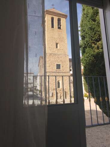 Through a window with sheer curtains, an old stone bell tower and a tall green cypress tree stand outside under a bright blue sky. Peaceful scene.