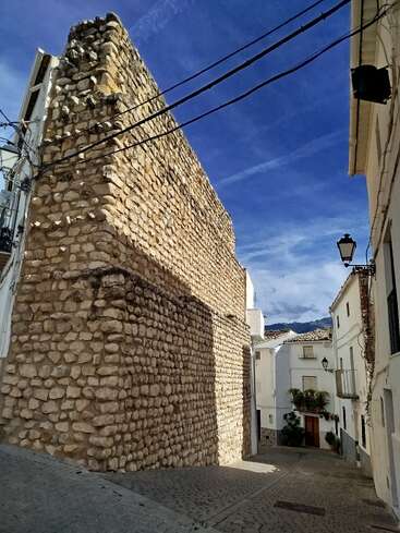 A narrow cobblestone street features old white houses and a prominent ancient stone wall, with clear blue skies overhead and mountains visible in the background.