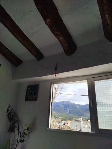 The image shows a white-walled room with dark wooden ceiling beams. A window reveals rooftops, houses, and distant mountains under a partly cloudy sky, creating a peaceful scene.