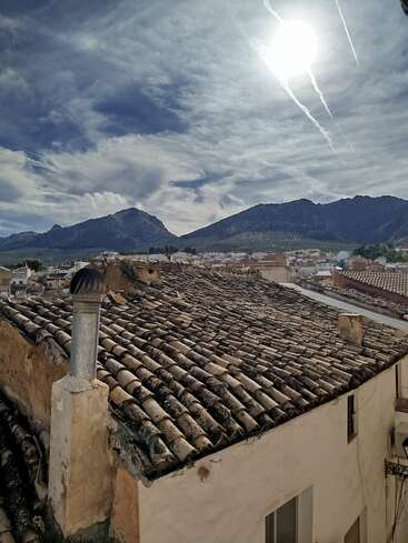 Old terracotta tiled rooftops in a rustic town, mountains in the background, under a dramatic sky with sun and contrails, creating a peaceful, picturesque atmosphere.