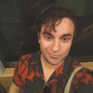 A man with wavy dark hair takes a selfie, wearing a red and black patterned shirt and sunglasses on his chest, seated on public transport.