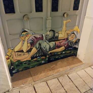 Eine gemalte Tür zeigt zwei Frauen, die im Gras liegen, eine träumt, die andere ruht sich aus. Helle Farben und ein skurriler Stil schaffen eine lebendige, verspielte Szene.