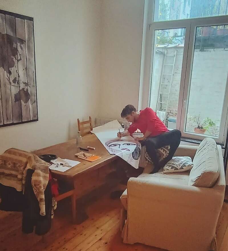 Eine Person in einem roten Hemd sitzt auf einer Couch, über einen Tisch gelehnt, und zeichnet ein großes Porträt. Der Raum ist gemütlich mit natürlichem Licht.