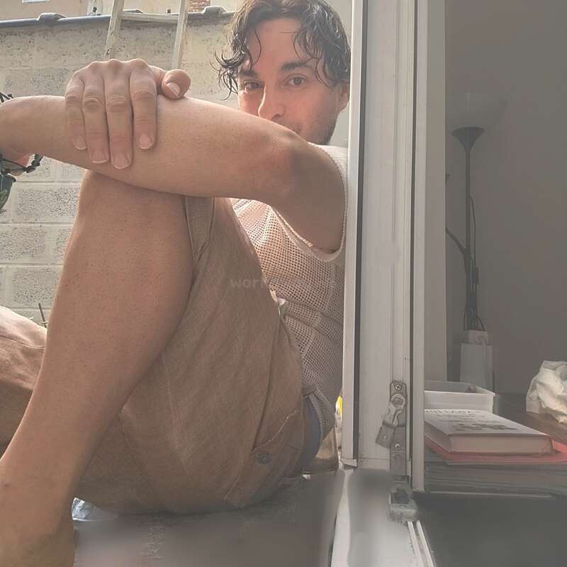 A man sits barefoot in a sunlit window, hugging his knees. He gazes outside, relaxed. Books, a lamp, and a soft indoor setting are visible.