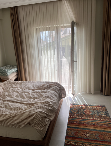 A luz do sol passa por cortinas transparentes em um quarto aconchegante com uma cama desfeita, tapete estampado e uma janela que dá para uma pequena varanda ou área externa.