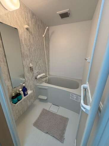 This is a compact, modern Japanese bathroom with a deep soaking tub, hand shower, small mirror, toiletries shelf, gray bath mat, and minimalistic design.