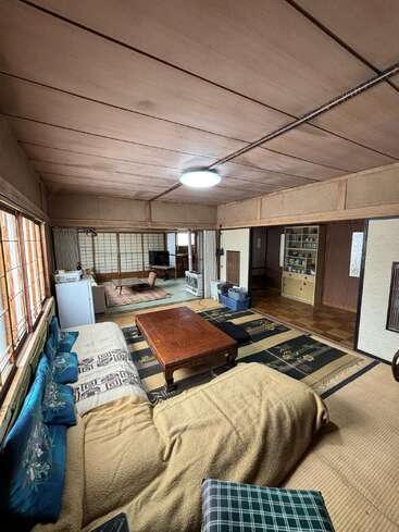 This image shows a traditional Japanese living room with tatami mats, futons, a low wooden table, shoji screens, cushions, minimal furniture, and natural light from windows.