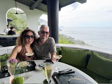 A smiling couple sits at a stylish outdoor restaurant table, enjoying cocktails with a breathtaking ocean view and lush green hills in the background, looking relaxed.