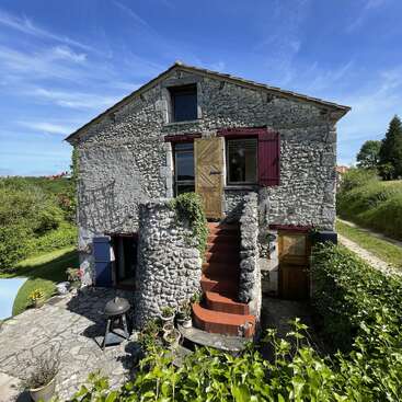 L'image représente une charmante maison en pierre avec une porte et un escalier en bois, entourée d'une végétation luxuriante et d'un ciel bleu serein par une journée ensoleillée.