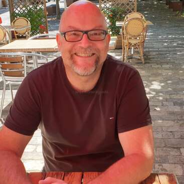 Un hombre sonriente con gafas y barba se sienta en una mesa de madera al aire libre en un café iluminado por el sol. Sillas vacías y vegetación rodean la agradable escena.