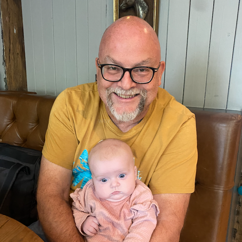 A smiling, bearded man wearing glasses and a yellow shirt is sitting on a brown sofa, lovingly holding a baby dressed in a pink outfit.