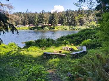 L'image représente une scène de lac sereine avec une verdure luxuriante, avec un foyer et des bancs, entourée d'arbres et d'une maison à l'arrière-plan, par une journée ensoleillée.