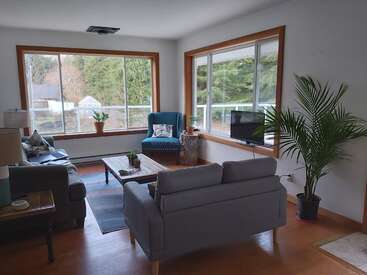 L'image représente un salon confortable avec deux fenêtres, comprenant un canapé, un fauteuil, une table basse, une télévision et des plantes, dégageant une atmosphère chaleureuse et accueillante.