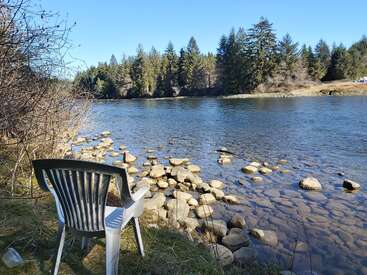 L'image représente une scène sereine au bord d'une rivière avec une chaise en plastique blanche face à l'eau, entourée de rochers et d'arbres sous un ciel bleu clair.