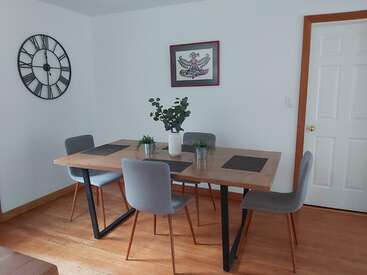 L'image représente une salle à manger avec une table en bois, quatre chaises et une grande horloge sur le mur, avec une plante et une œuvre d'art au centre.