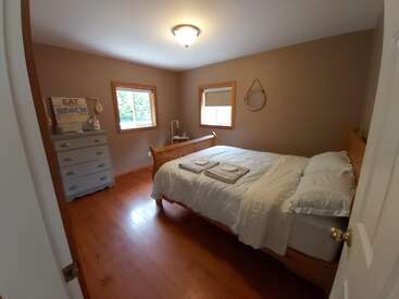 L'image représente une chambre à coucher confortable avec un lit double, des draps blancs et une commode, dotée d'une fenêtre et d'une porte, avec une atmosphère sereine et une lumière naturelle.