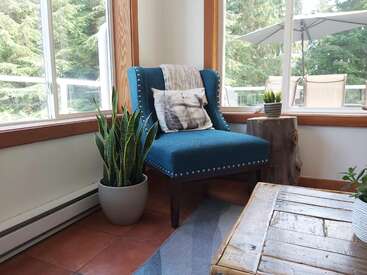 L'image représente une pièce confortable avec une chaise bleue, une table basse en bois et des plantes en pot, sur fond de fenêtres et d'un patio extérieur.