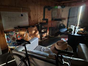 A cozy wooden cabin interior with a bicycle, telescope, wood stove, colorful chair, table with food, hanging helmets, sofa, hats, and sunlight streaming in.