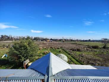 A corrugated metal roof in the foreground overlooks a vast, green countryside with scattered trees, fences, blue sky, and a few distant buildings under sunlight.
