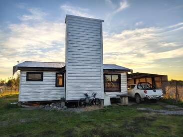 A small, modern white house stands on grass with an unusual tall center chimney. A parked white pickup truck, bike, and wooden bench complete the scene.