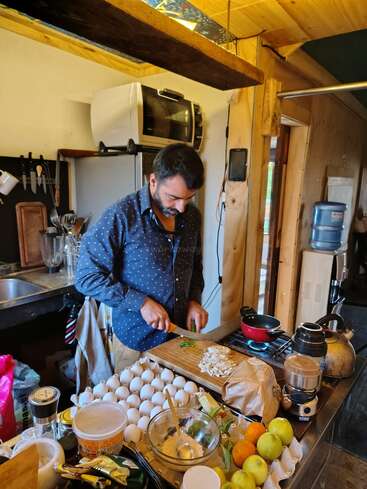A man is chopping vegetables in a cozy, rustic kitchen. Eggs, fruits, and kitchen utensils clutter the counter, while a kettle heats on the stove nearby.