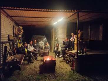 A group of friends gather under a corrugated roof, sitting around a small fire pit. They smile, enjoy drinks, and relax in a rustic setting.