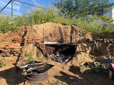 This image shows an outdoor cooking setup with a fire pit built into a dirt embankment, surrounded by stacked firewood, cooking tools, greenery, and bright sunlight.