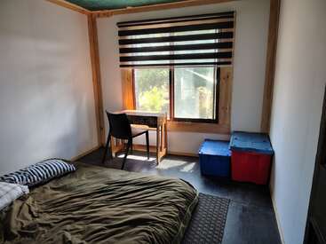 Esta imagen muestra un dormitorio sencillo con una cama, almohadas a rayas, un pequeño escritorio y una silla junto a la ventana, y dos cubos de almacenaje en el suelo.