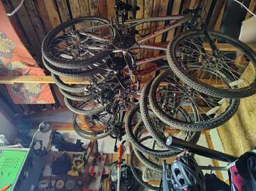 Varias bicicletas cuelgan boca abajo de un techo de madera en un garaje o taller desordenado, rodeadas de herramientas, cascos, luces, armarios y material.