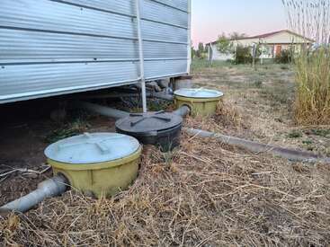 Two large yellow and one black plastic tanks connected with pipes sit beside a corrugated metal building. The ground is dry with sparse grass, house in background.