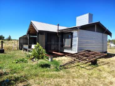 A small, rustic wooden house with a metal roof stands in a grassy, rural area. A metal trailer sits in front, under a bright blue sky.
