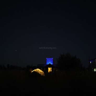 Uma cena noturna escura apresenta uma casa com luzes azuis iluminando uma torre. Uma tenda brilhante fica nas proximidades, cercada por silhuetas de árvores e grama.