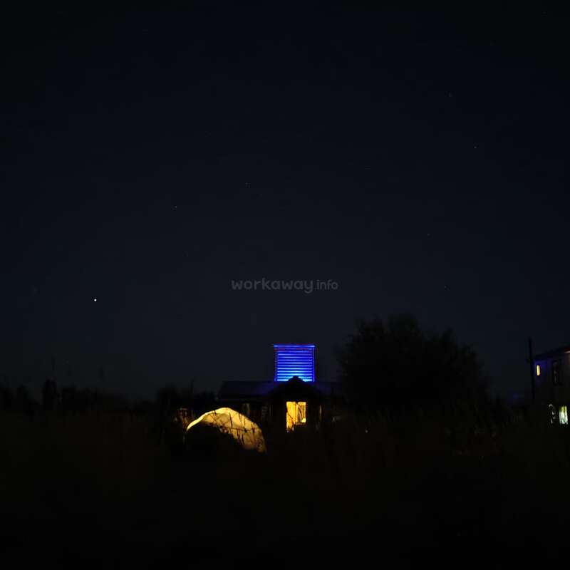 A dark night scene features a house with blue lights illuminating a tower. A glowing tent sits nearby, surrounded by silhouettes of trees and grass.